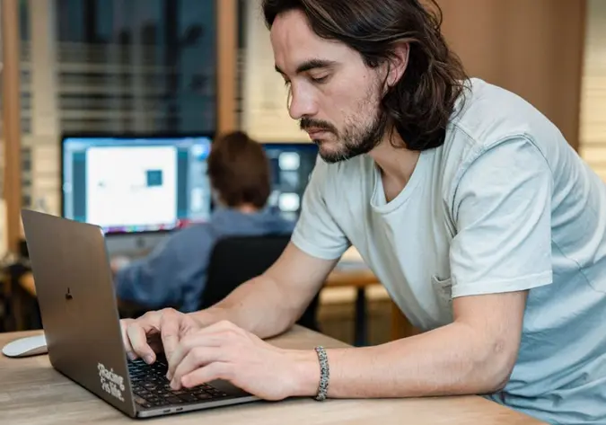 Foto von zwei Personen an Computern, eine im Vordergrund, eine im Hintergrund mit zwei Monitoren.